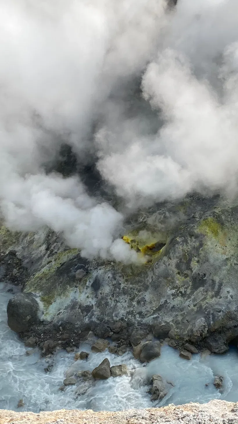 Steam lifting off sulphur-stained rock into pale water, a planet still busy with its own making.